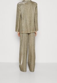 Traje dorado brillante que presenta una chaqueta de manga larga ajustada y pantalones de pierna ancha, ambos con un brillo metálico y una superficie texturizada.