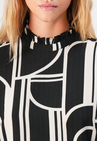 Primo piano di una donna che indossa un top a fantasia geometrica bianco e nero con un piccolo colletto a ruches e labbra lucide in evidenza.