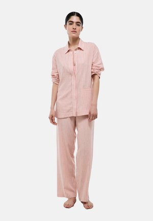 Femme debout pieds nus portant un pyjama rose ample avec de fines rayures verticales rouges, une chemise boutonnée à manches longues et un pantalon à jambes larges.