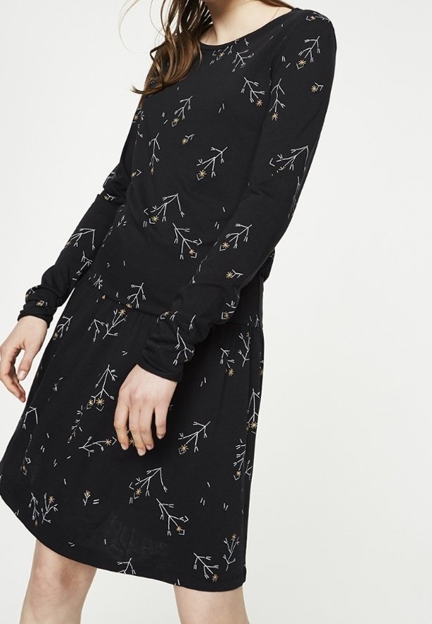 Femme portant une robe noire à manches longues avec un motif floral subtil composé de petites branches et fleurs blanches et beige.