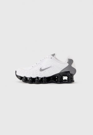 Chaussure de course Nike blanche et grise avec une semelle noire segmentée, présentant une tige texturée et le logo Nike sur le côté.