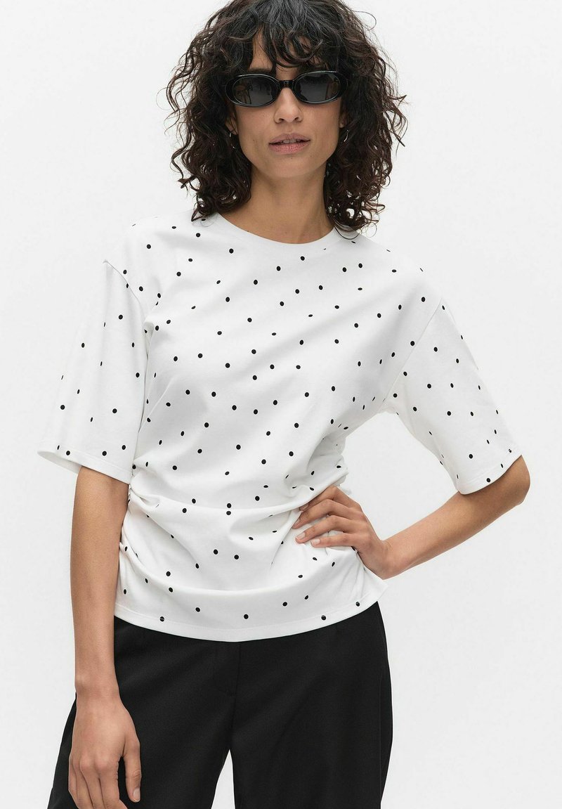 Nainen, jolla on kiharat hiukset ja mustat aurinkolasit, valkoinen lyhythihainen paita, jossa mustia pilkkuja, sekä mustat housut, poseeraa käsi lantiolla.