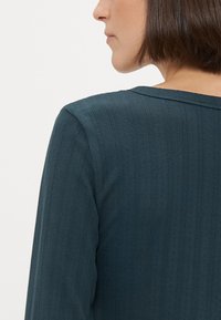 Ciemnozielony sweter z dzianiny, z pionowym prążkowanym wzorem, z okrągłym dekoltem i dopasowanym krój. Tył jest częściowo widoczny.