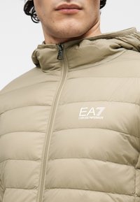 Jachetă matlasată bej, cu textură quiltată, glugă și fermoar frontal. Prezintă logo-ul EA7 Emporio Armani alb pe partea stângă a pieptului.