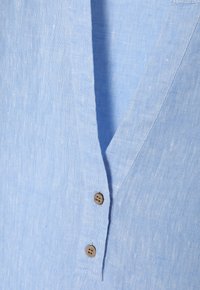 Camicia in lino azzurro chiaro con scollo a V, due bottoni marroni e una texture morbidissima, adatta per un abbigliamento casual.