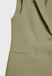 Gilet croisé en vert olive clair, fabriqué en tissu lisse. Comprend deux boutons à l'avant et une poche latérale avec une ouverture étroite.