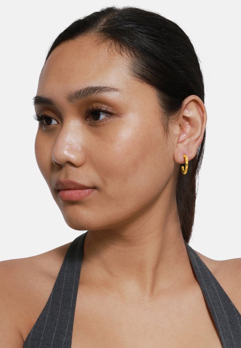 Goldene Creolen getragen, mit einer glatten, polierten Oberfläche. Das Modell hat einen mittleren Hautton und dunkles Haar, mit einem neutralen Ausdruck.