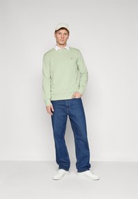 Lichtgroene trui van zacht materiaal, gecombineerd met een wit overhemd met kraag, donkerblauwe jeans en witte sneakers. Een strakke pet maakt de look compleet.
