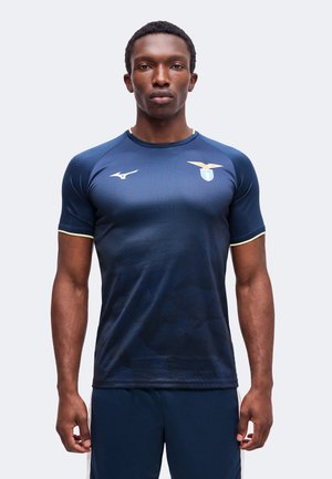 Uomo che indossa una maglietta sportiva blu scuro con un logo bianco sul petto e rifiniture gialle sulle maniche, in piedi davanti a uno sfondo semplice.
