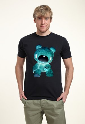 Ung mand med lyst hår iført en sort T-shirt med et stort blågrønt billede af en bamse, kombineret med lyse bukser.