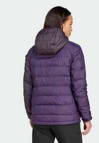 Frau in einer lila und dunkelgrauen gepolsterten Kapuzenjacke, die von hinten zu sehen ist, mit entspannten Händen an den Seiten vor einem einfarbigen Hintergrund.