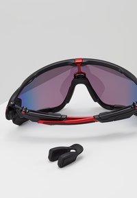 Occhiali da sole con montatura nera e lenti a gradiente viola. Le caratteristiche includono dettagli rossi, lati regolabili e un para-naso removibile.