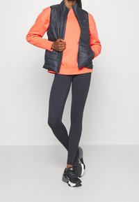 Mustat pehmeät liivit oranssin collegepaidan päällä, yhdistettynä tummaan leggingseen. Asusteena mustat urheilukengät, joissa on valkoisia yksityiskohtia.