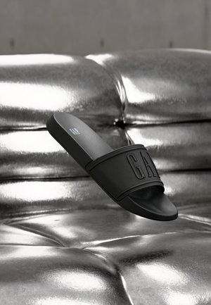 Sandale slide noire avec une large lanière et le logo "GAP", posée sur une surface capitonnée métallique brillante.