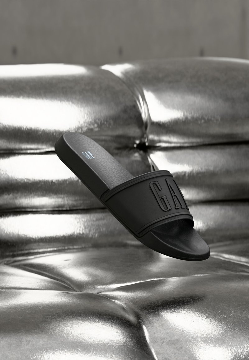 Sandale slide noire avec une large lanière et le logo "GAP", posée sur une surface capitonnée métallique brillante.