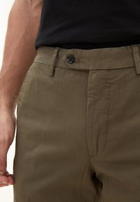 I pantaloni in cotone verde oliva presentano una chiusura a bottone, tasche laterali e una texture liscia. La vita è piatta con passanti per cintura.