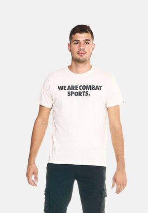 Jonge man met kort haar, die een wit t-shirt draagt met de tekst "WE ARE COMBAT SPORTS" en zwarte cargobroek, staand tegen een effen achtergrond.
