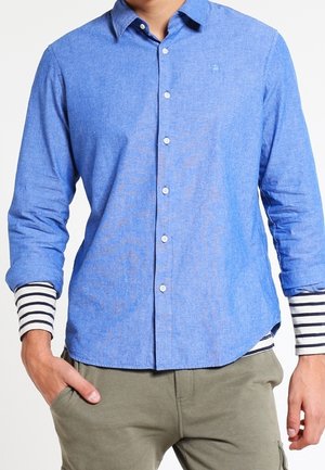 Homme portant une chemise bleue à boutons sur un t-shirt à manches longues rayé noir et blanc, les mains partiellement dans les poches d'un pantalon vert olive.