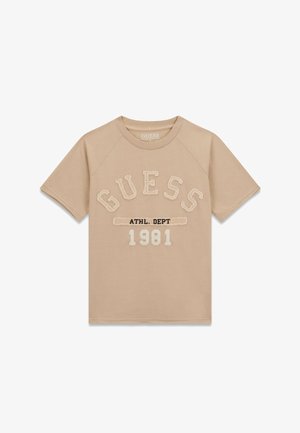 T-shirt beige a maniche corte con la scritta "GUESS" in grandi lettere, "ATHL. DEPT" in caratteri più piccoli e "1981" stampato sotto, sul davanti.