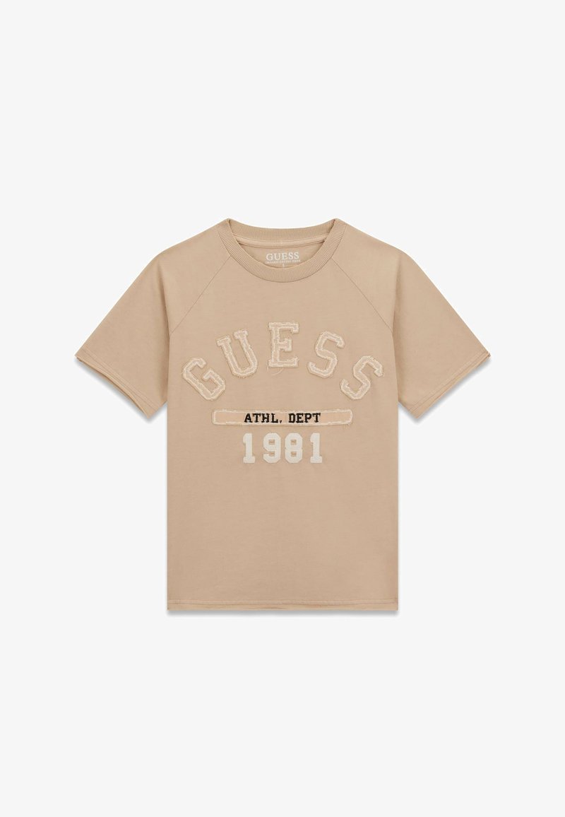 T-shirt beige à manches courtes avec "GUESS" en grandes lettres, "ATHL. DEPT" en texte plus petit, et "1981" imprimé en dessous sur le devant.