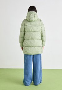 Jachetă puffer verde deschis cu glugă, cu cusături orizontale și o croială lungă, asociată cu blugi evazați albaștri.