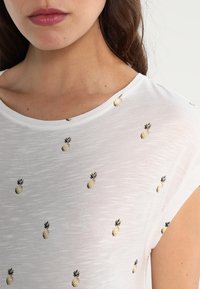 Camicia bianca a maniche corte realizzata in un tessuto leggero e testurizzato, con piccoli motivi di ananas ricamati in giallo e verde.