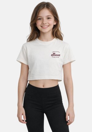 Fille souriante portant un t-shirt court blanc avec le logo « ellesse » et des leggings noirs taille haute, debout devant un fond uni.