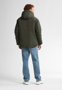 Donkergroene pufferjack met capuchon, met een gladde textuur. Draagt zich met lichtblauwe spijkerbroek en witte sneakers. Achteraanzicht getoond.