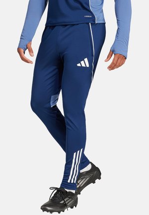 Marineblauwe sportbroeken van stretchstof met witte drie-strepenaccenten, een zijzak en een taps toelopende pasvorm, gecombineerd met zwarte voetbalschoenen.
