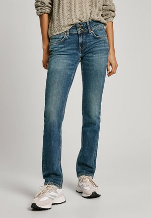 Blaue Denim-Jeans mit geradem Schnitt, mittlerer Waschung, dezenten Verblassungen und doppeltem Knopfverschluss, kombiniert mit hellen Sneakers.