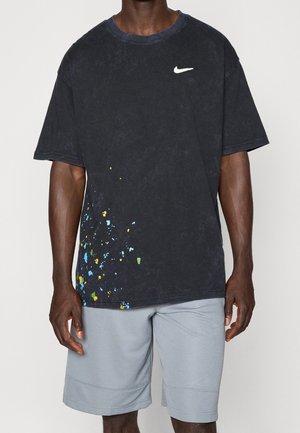 Hombre vestido con camiseta negra Nike con salpicaduras de pintura multicolor en la parte inferior izquierda y pantalones cortos gris claro, de pie frente a un fondo liso.