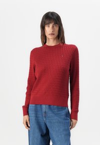 Suéter de punto rojo con textura acanalada, cuello redondo y mangas largas, combinado con jeans de mezclilla azules. Pequeño detalle del logo en el pecho.