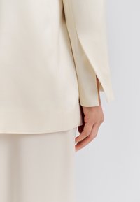 Béžový saténový blejzer s rovný strihom a dlhým rukávom, ukazujúci bočný rozparok. Viditeľná ruka spočíva na zodpovedajúcej béžovej saténovej sukni.