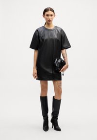 Vestido de couro preto de mangas curtas com decote redondo, combinado com botas pretas até ao joelho e uma pequena bolsa preta com cordão. Textura suave.