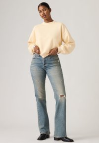 Γυναίκα με κρεμ φαρδιά φούτερ, φθαρμένα ανοιχτό μπλε τζιν με σκίσιμο στο γόνατο και μαύρα μποτάκια αστραγάλου, που στέκεται μπροστά σε απλό φόντο.