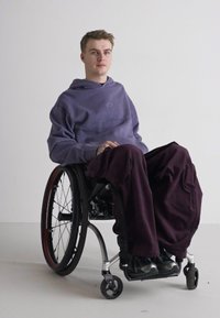 Scaun cu rotile negru cu accente argintii, o persoană îmbrăcată cu un hanorac mov și pantaloni largi de culoare închisă, așezată pe un fundal alb simplu.