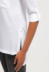 Vit blus med en frampocket, uppvikta ärmar, sidokilar och en etikett vid fållen. Har ett mjukt, lätt tyg.