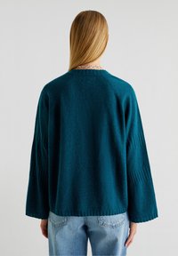 Pulover oversized teal cu mâneci late, detalii cu un model rib și un finisaj moale, texturat. Purtați peste blugi albaștri.