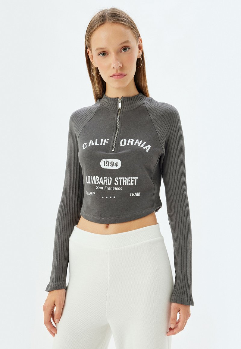 Grijze cropped sweatshirt met een hoge kraag en rits, geribbelde textuur, met witte grafische tekst en accenten. Gecombineerd met witte broek.