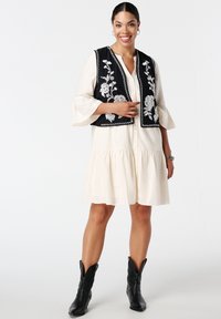 Zwarte geborduurde vest met bloemmotieven over een lichtbeige jurk met een uitlopende rok en lange, klokvormige mouwen. Zwarte cowboylaarzen.