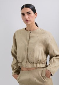 Chaqueta beige cortada con un escote redondeado, adornada con patrones de olas plateados y dobladillo elástico. Tejido suave con un ligero brillo.