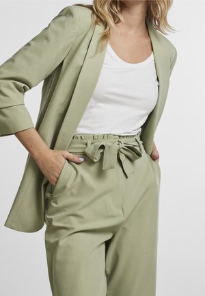 Vrouw draagt lichtgroene blazer en bijpassende hoog getailleerde broek met strikriem, wit shirt met ronde hals, handen in de zakken.