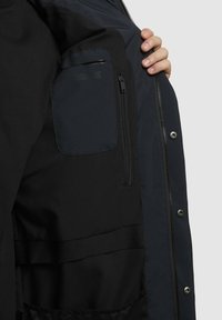 Fekete dzseki, bal oldalon cipzáras zsebbel. Az anyag sima textúrájú, kontrasztos sötétkék részletekkel.