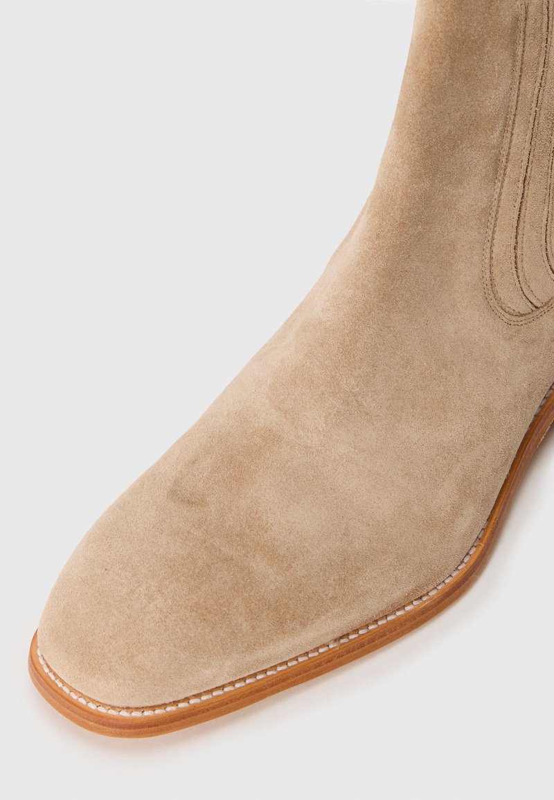 Bottines en daim beige avec une texture lisse, un bout arrondi et une semelle contrastante marron clair. Coutures subtiles le long des bords.