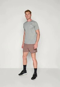 Ung mann som står i grå atletisk t-skjorte, brune shorts, svarte sokker og svarte løpetreningsko foran en ensfarget bakgrunn.