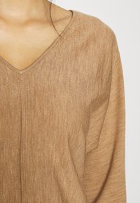 Maglione beige a scollo a V con un knit leggero e testurizzato. Presenta una lucentezza sottile ed è leggermente oversize, evidenziando un design rilassato.