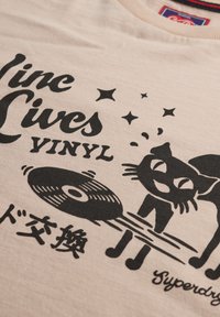 Beige bomuld T-shirt med sort grafik af en kat, der spiller en vinylplade, med teksten "Vinyl Lives" over og japanske tegn nedenunder.