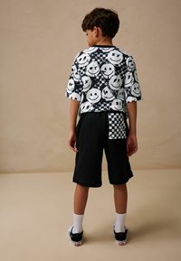 T-shirt à manches courtes noir et blanc avec motif de visage souriant, assorti à un short noir avec une poche à carreaux. Chaussettes blanches et baskets noires.