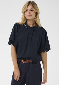 Haut bleu marine à manches courtes avec un col rond, détail froncé et coupe décontractée. Associé à un pantalon foncé et une ceinture marron.