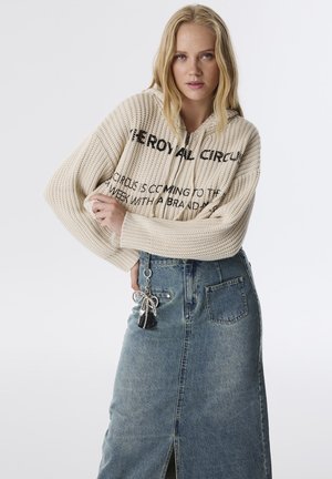 Beige Strickpullover mit schwarzem Text, kombiniert mit einem hochtaillierten Denim-Rock mit seitlichem Schlitz und einer dekorativen Schlüsselanhänger am Taschen.